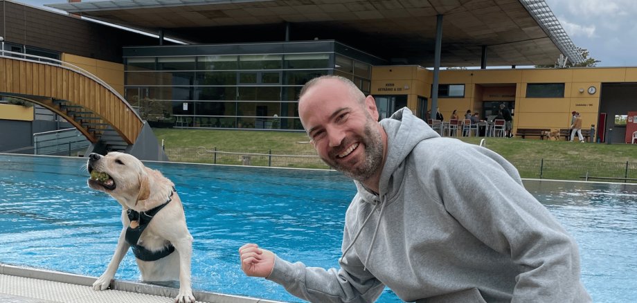 Hund in einem Schwimmbecken, daneben ein Mann sitzend am Beckenrand