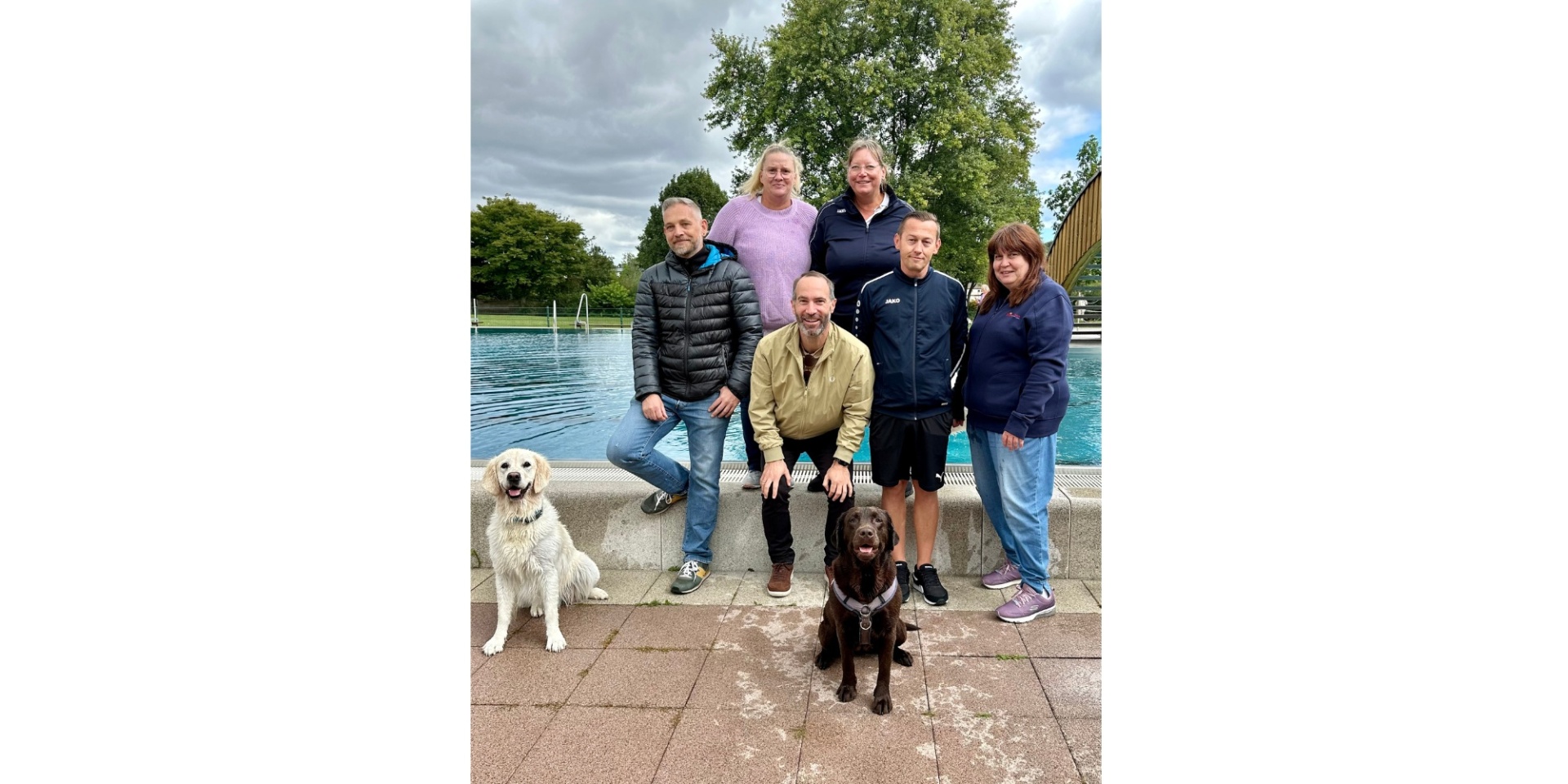 Mehrere Personen stehend mit zwei Hunden vor dem Beckenrand eines Freibades