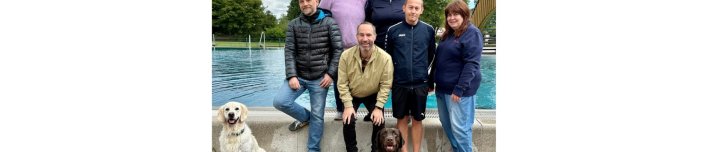 Mehrere Personen stehend mit zwei Hunden vor dem Beckenrand eines Freibades