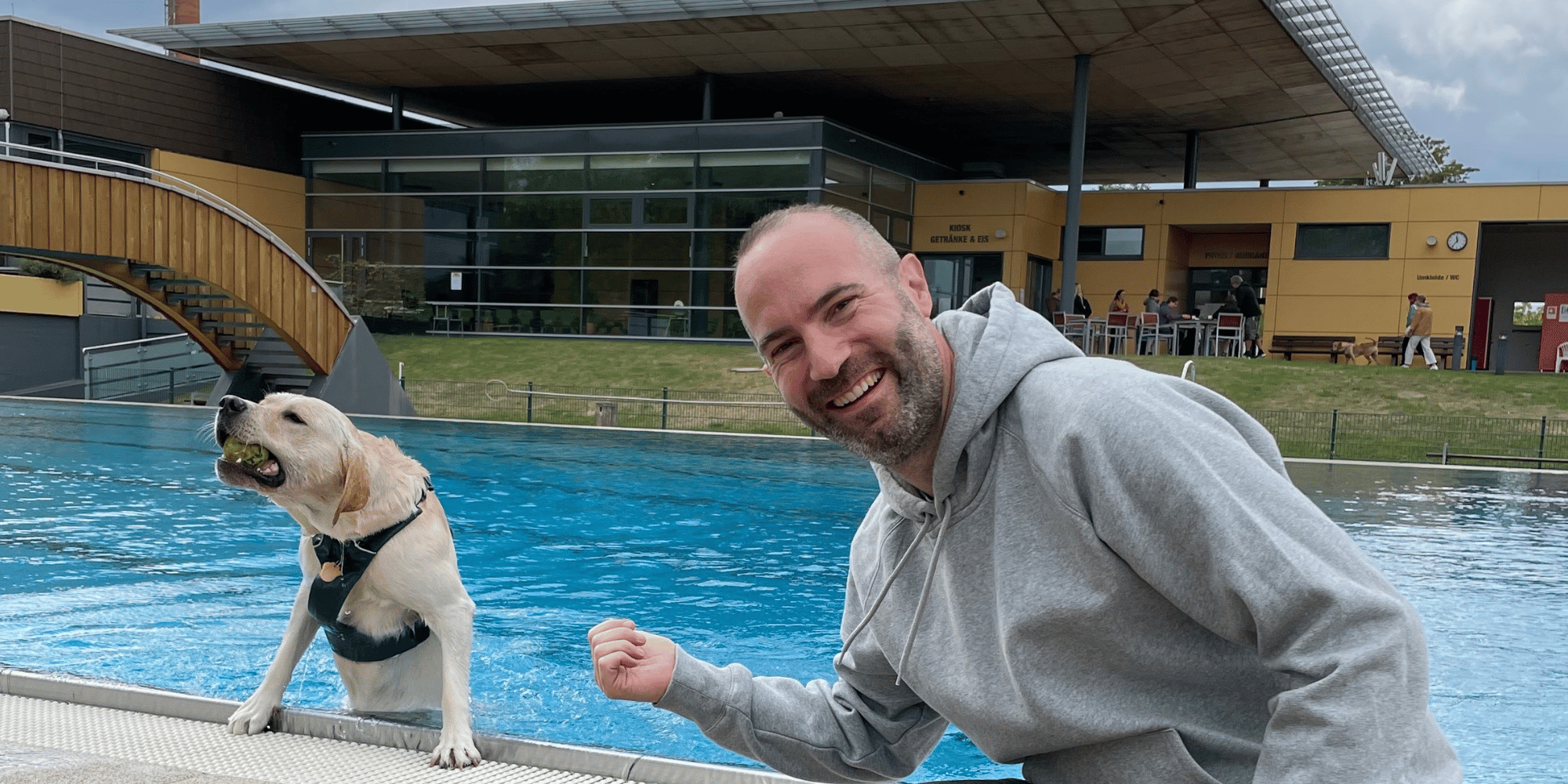Hund in einem Schwimmbecken, daneben ein Mann sitzend am Beckenrand